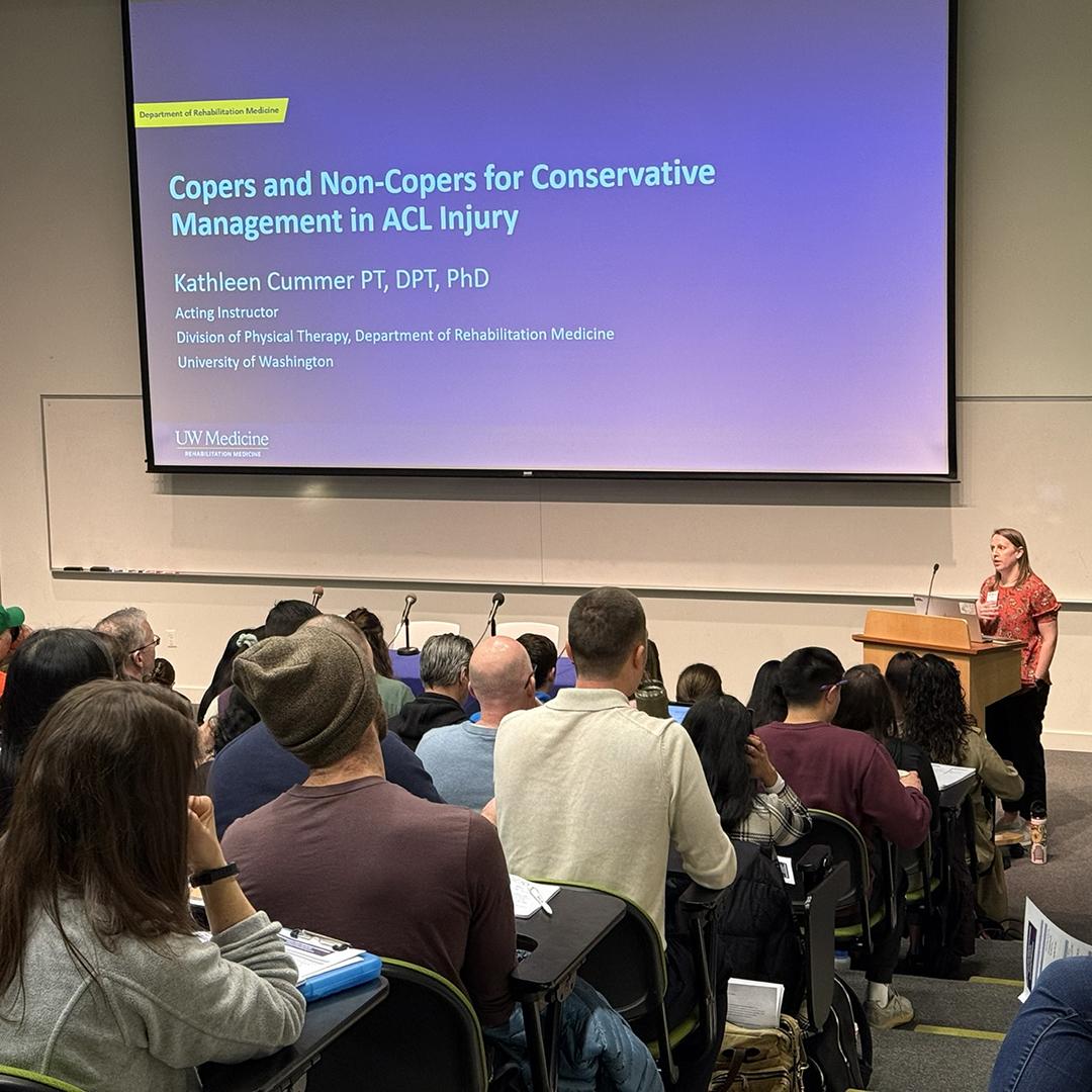 A woman speaks to crowd under a slide about copers and non-copers for conservative management in ACL injury. 