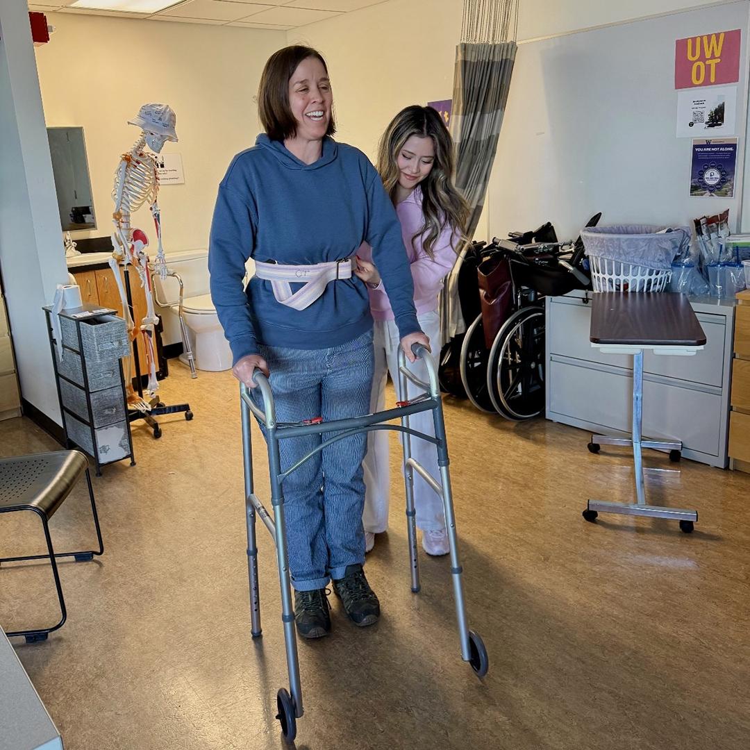 A woman in pink assists a woman in blue who is walking with a walker. 