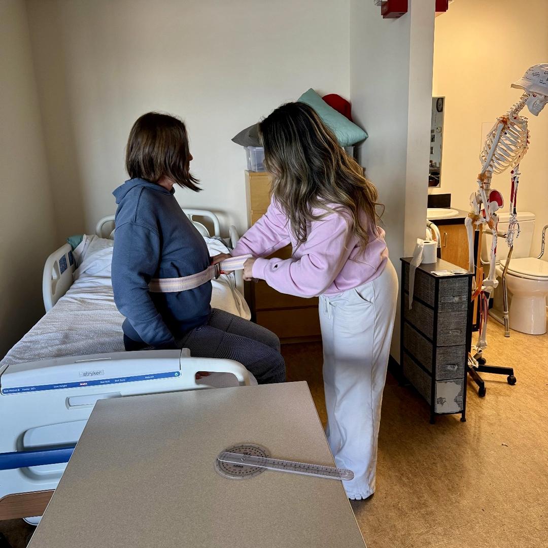 A woman in pink wraps a strap around a woman in blue seated on a bed. 