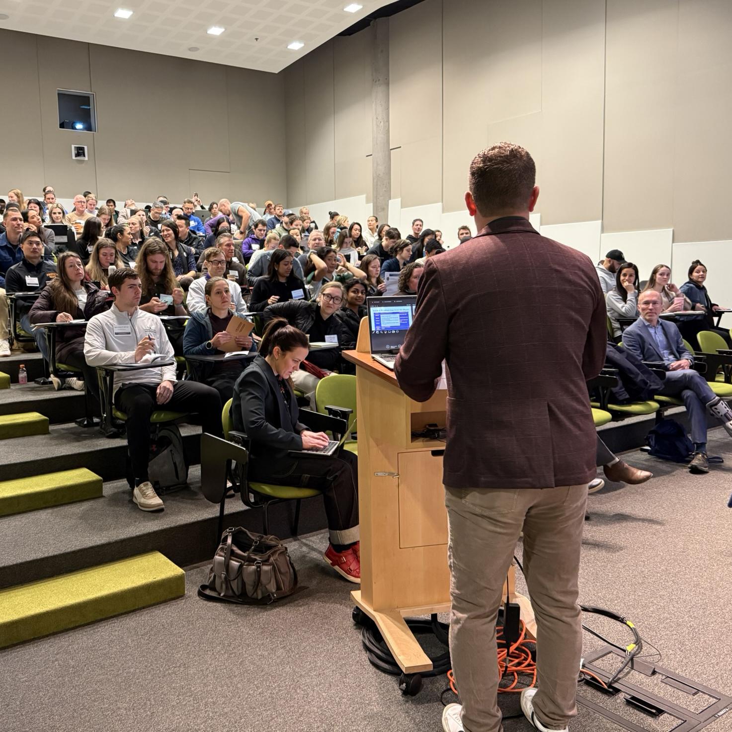 A man looks at a full classroom from behind a podium. 