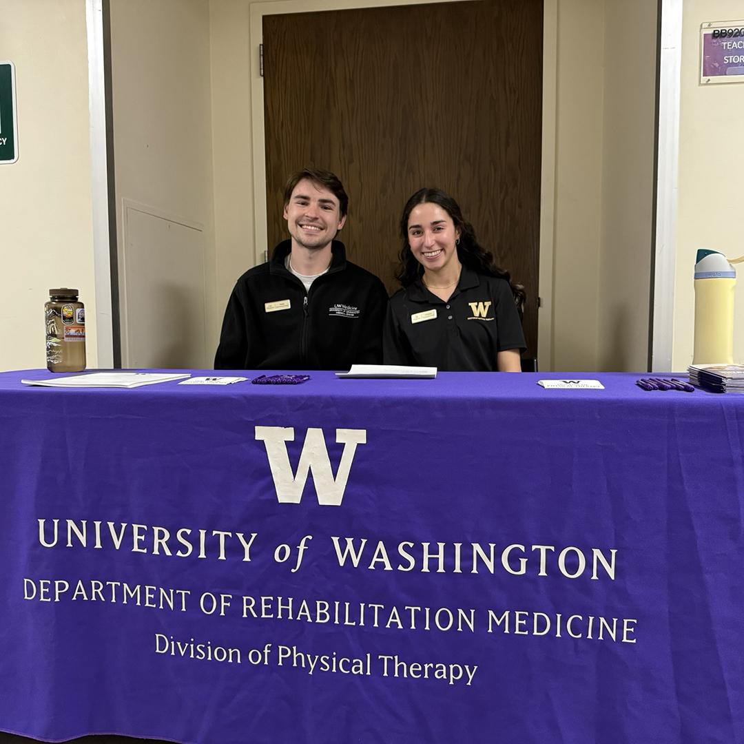 Two people smile from behind a table with a purple tablecloth. 