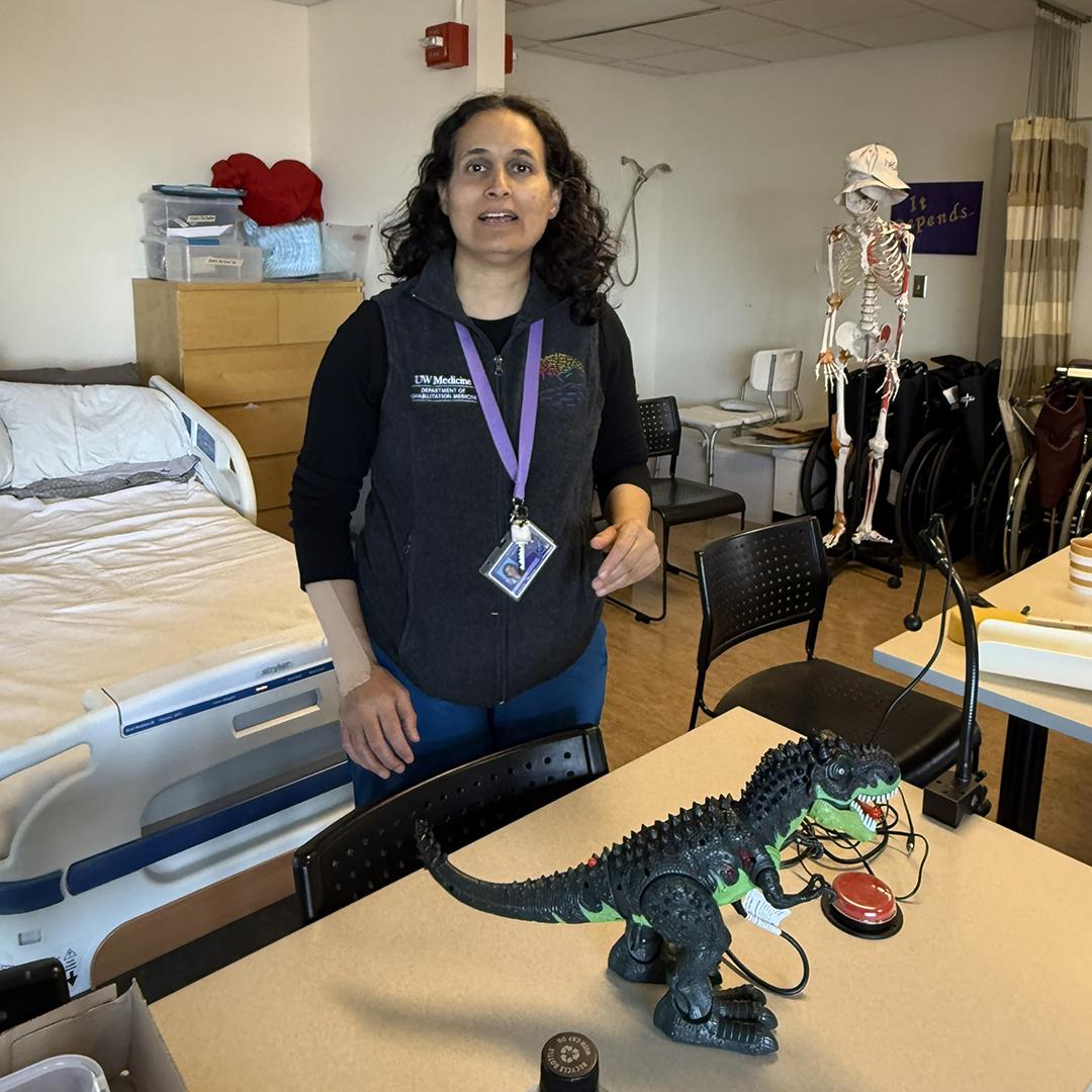 A woman stands next to a table where a toy dinosaur sits. 