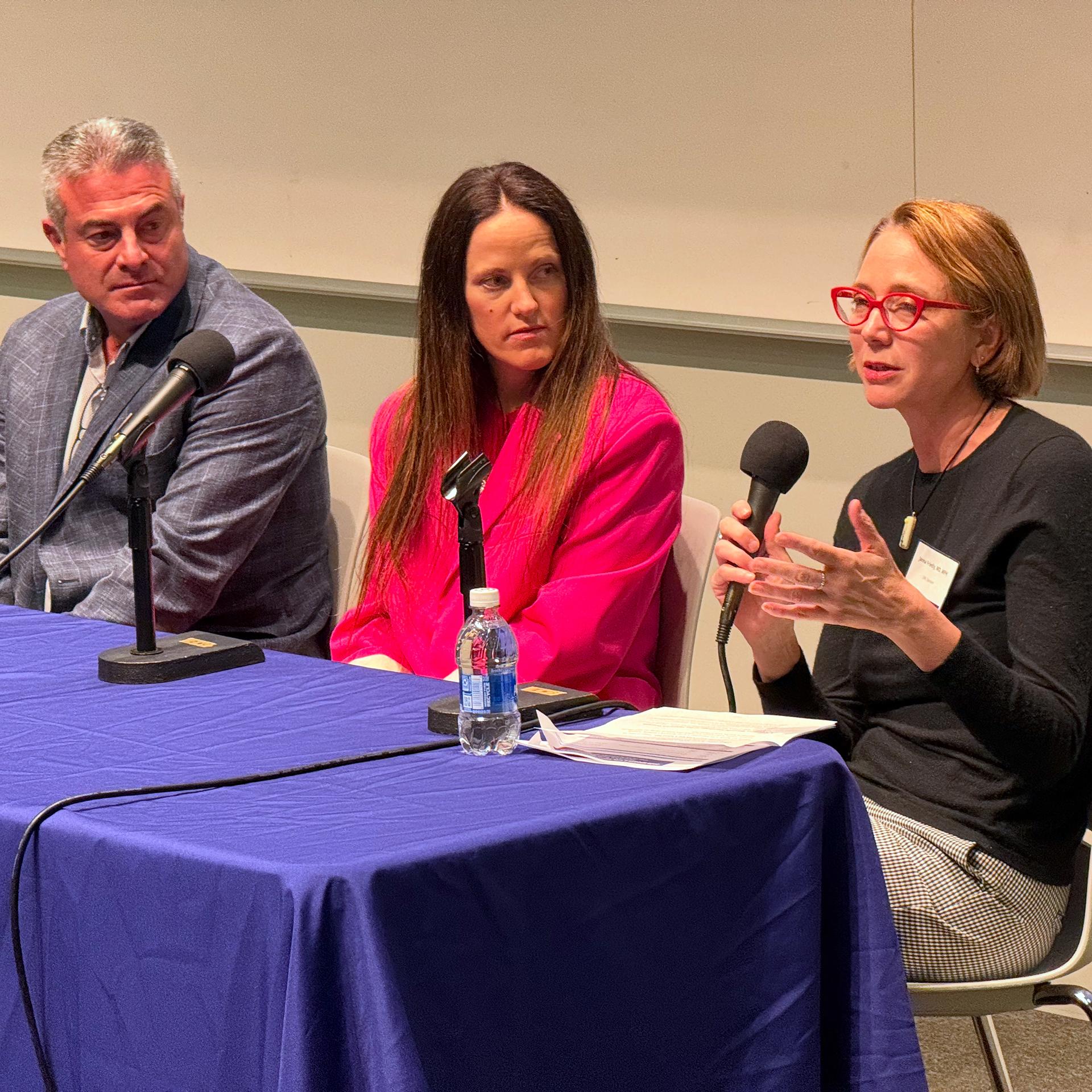 Three people sit at a table; one is speaking into a microphone. 