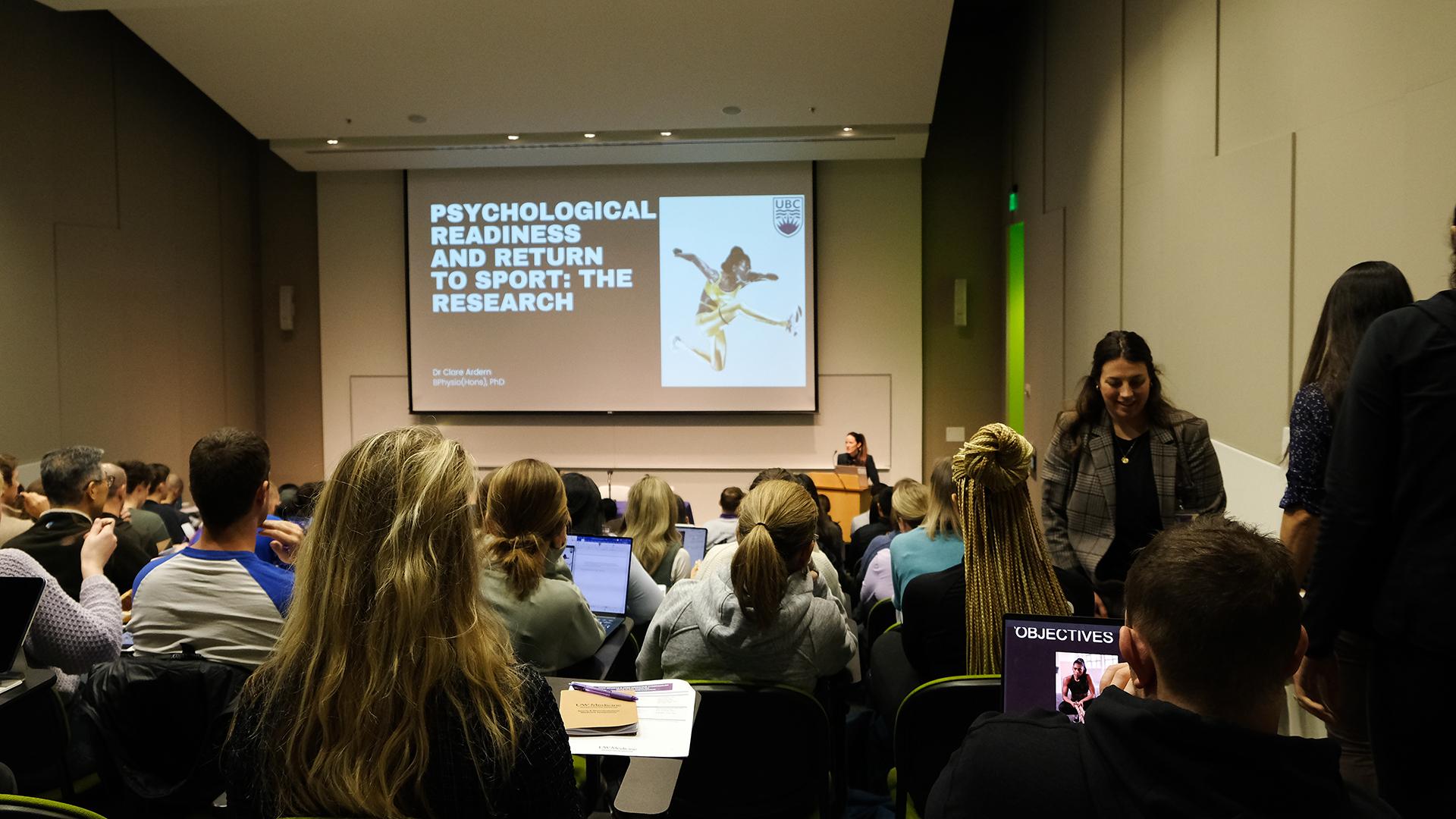 A woman speaks to a crowd under a slide about psychological readiness and return-to-sport.