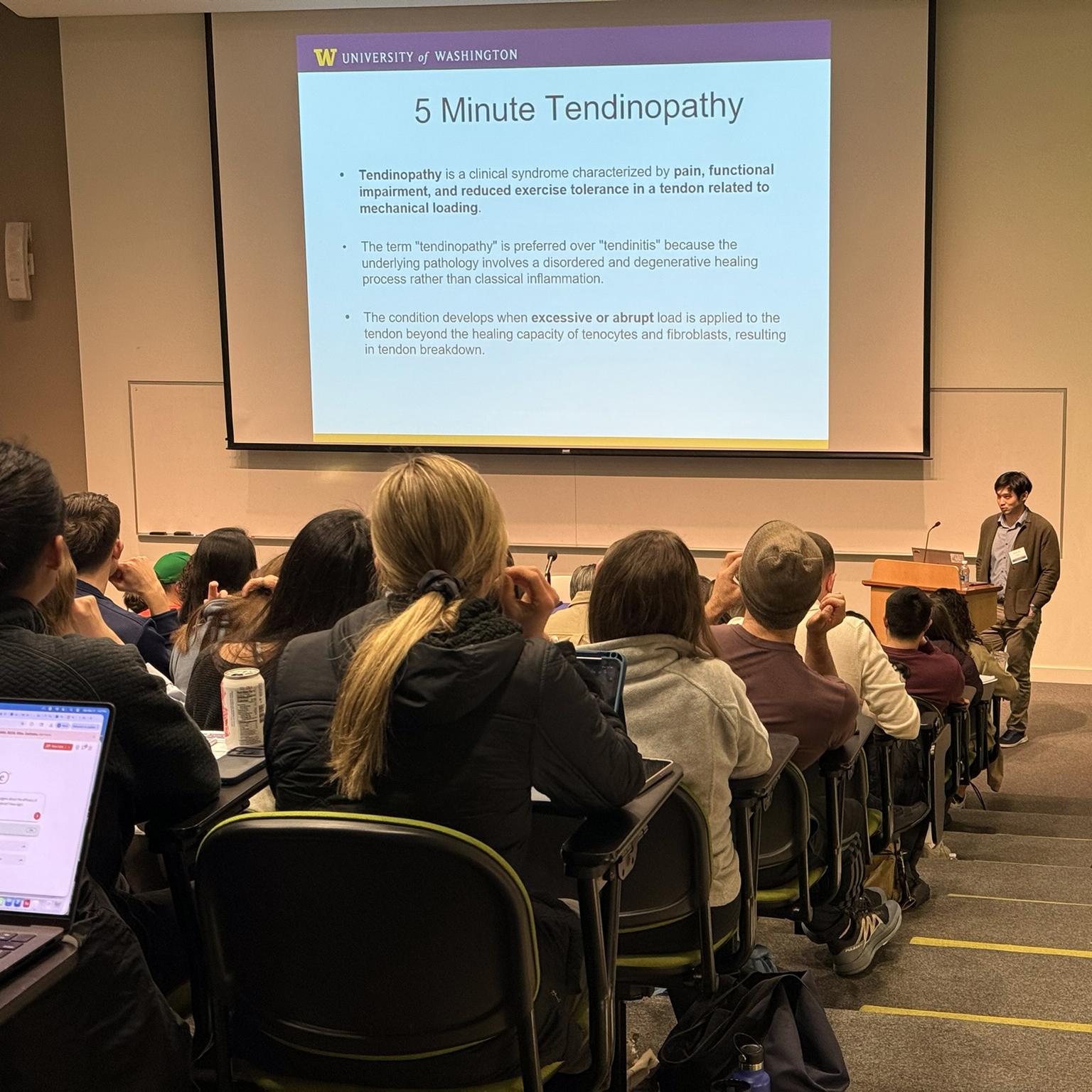 A crowd looks at a slide on 5 minute Tendinopathy. 