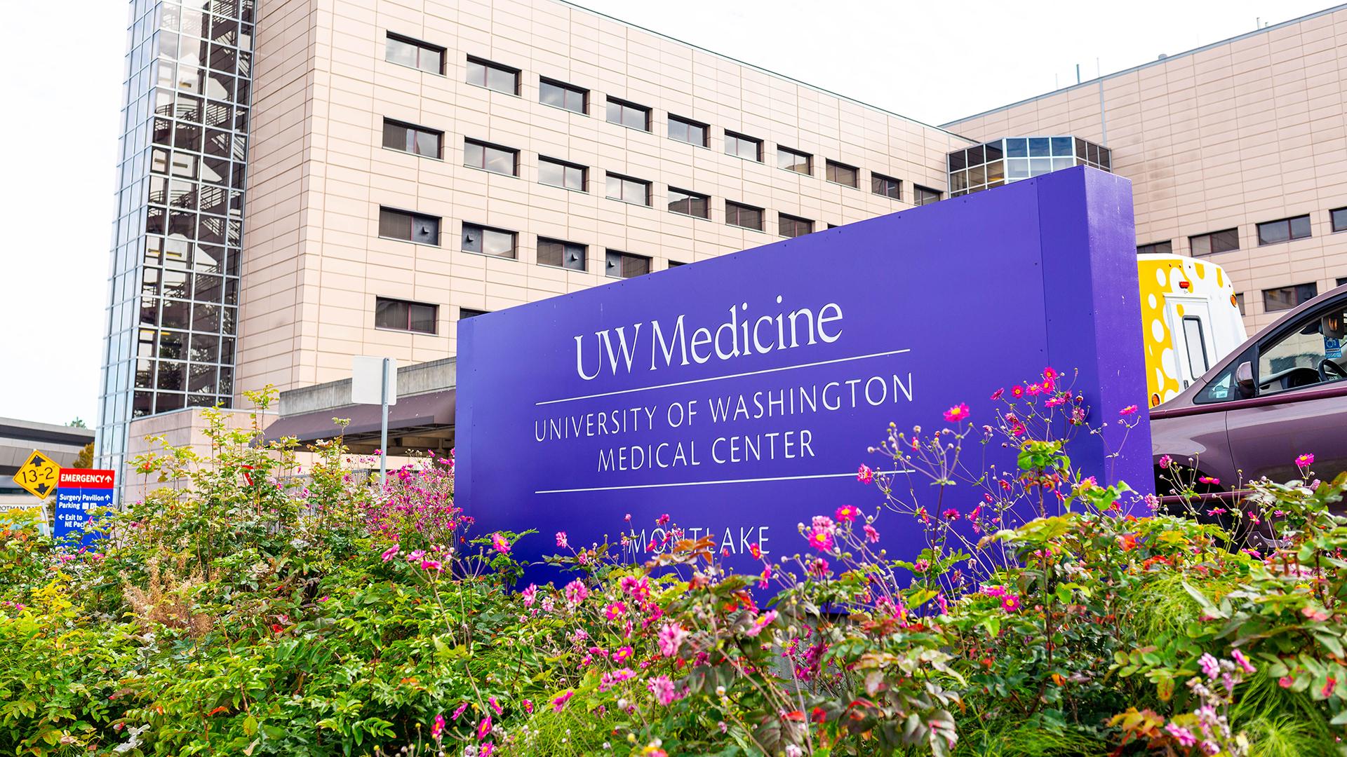 A purple sign for UW Medical Center – Montlake sits in a bed of flowers outside a large building.