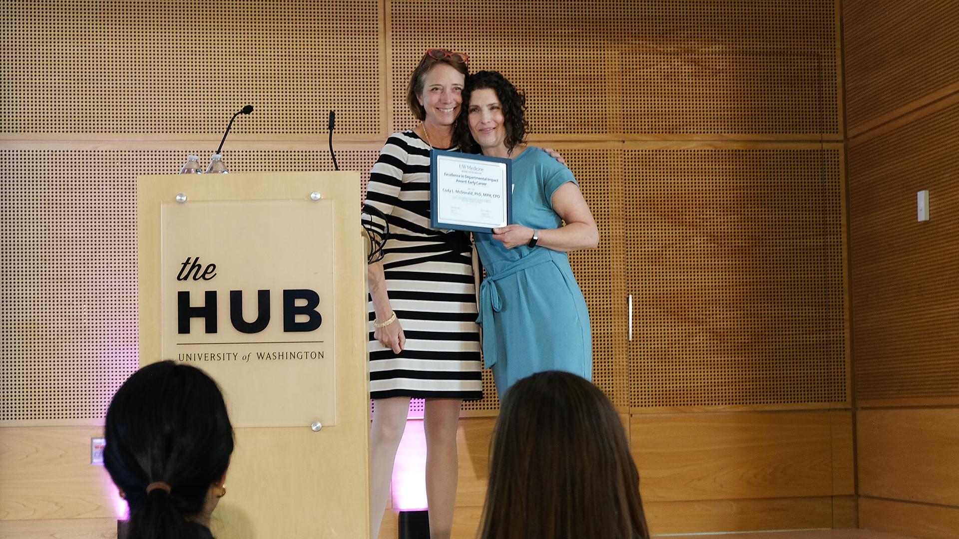 Two women smile on stage while one holds a certificate.