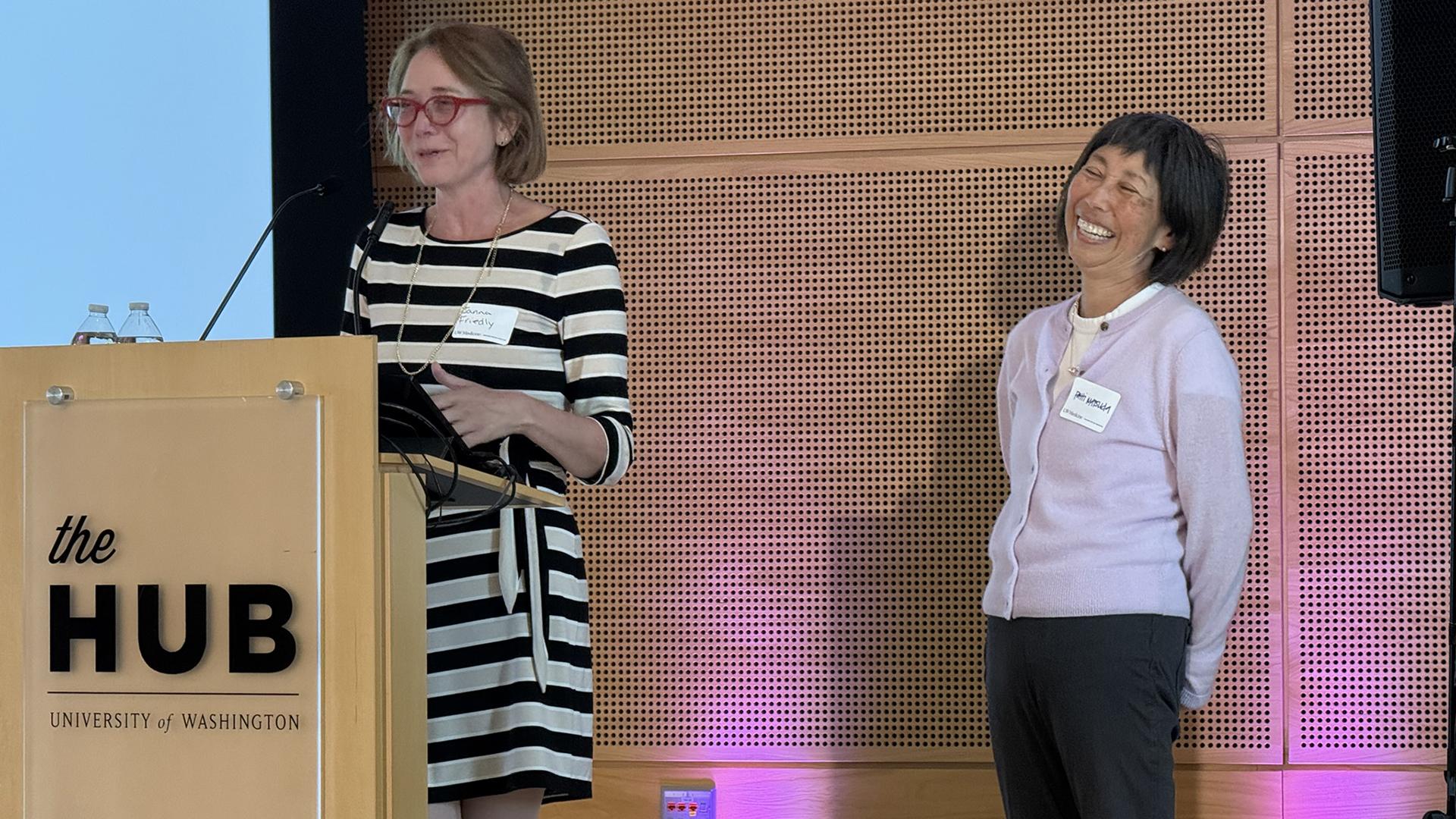 A woman speaks at a podium while another woman waits, laughing.