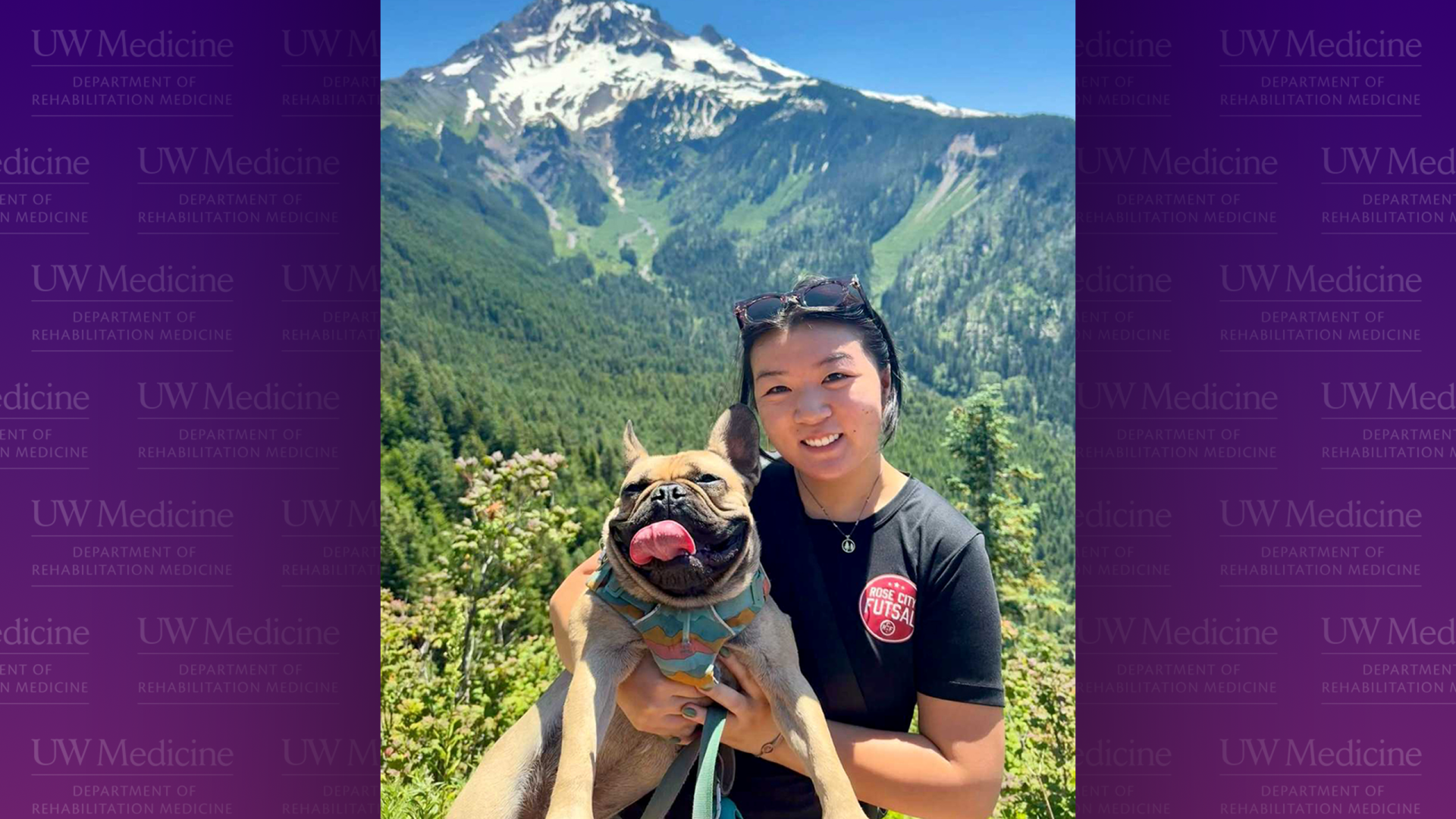 A woman holds a dog with a mountain rising behind her.